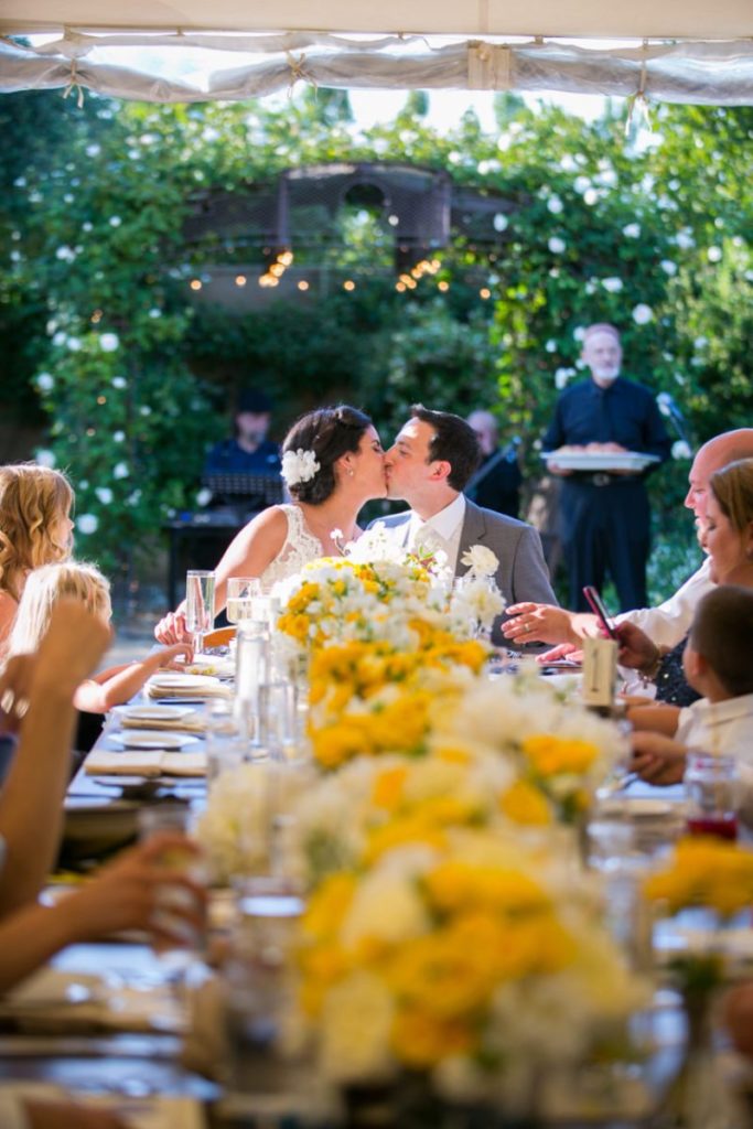 A breath-taking wine country wedding venue is features in this sweet photo of a bride and groom kissing at a table covered in yellow and white blossoms. Event by Blissful Events. Photo by Carmen Alvarez Photography.