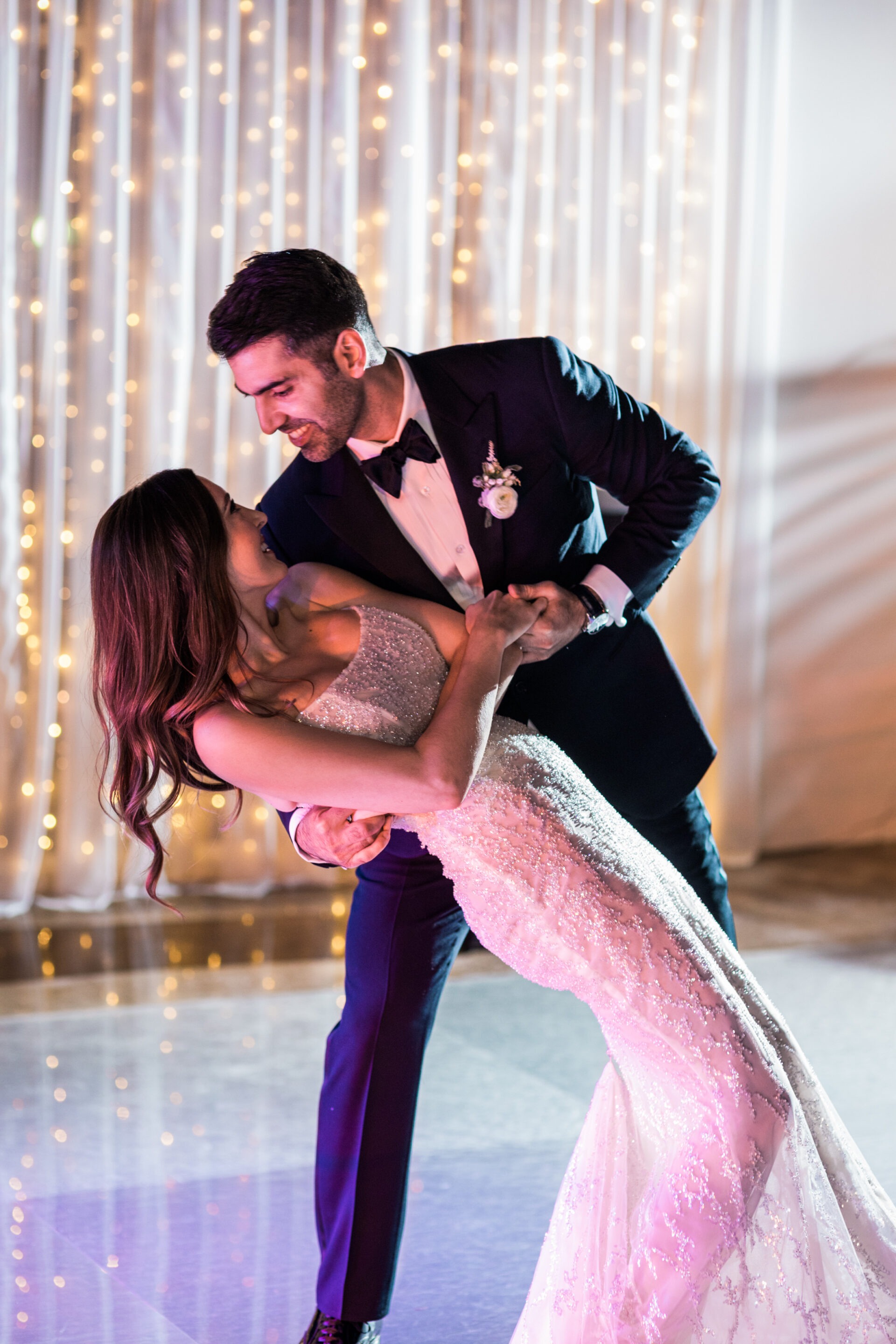 California DJ services can make any wedding an even more joyous event. A groom dips his bride on the dance floor with a curtain of twinkle lights in the background. 