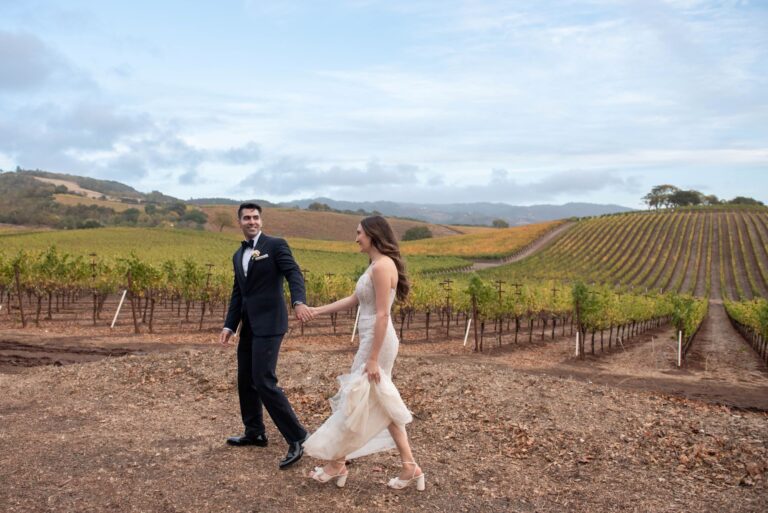 A newly wedded couple stroll on the grounds of Kunde Winery Wedding Venue & Wine Cave in front of rows of grape vines. Event by Blissful Events. Photo by Jessica Schmitt Photography.