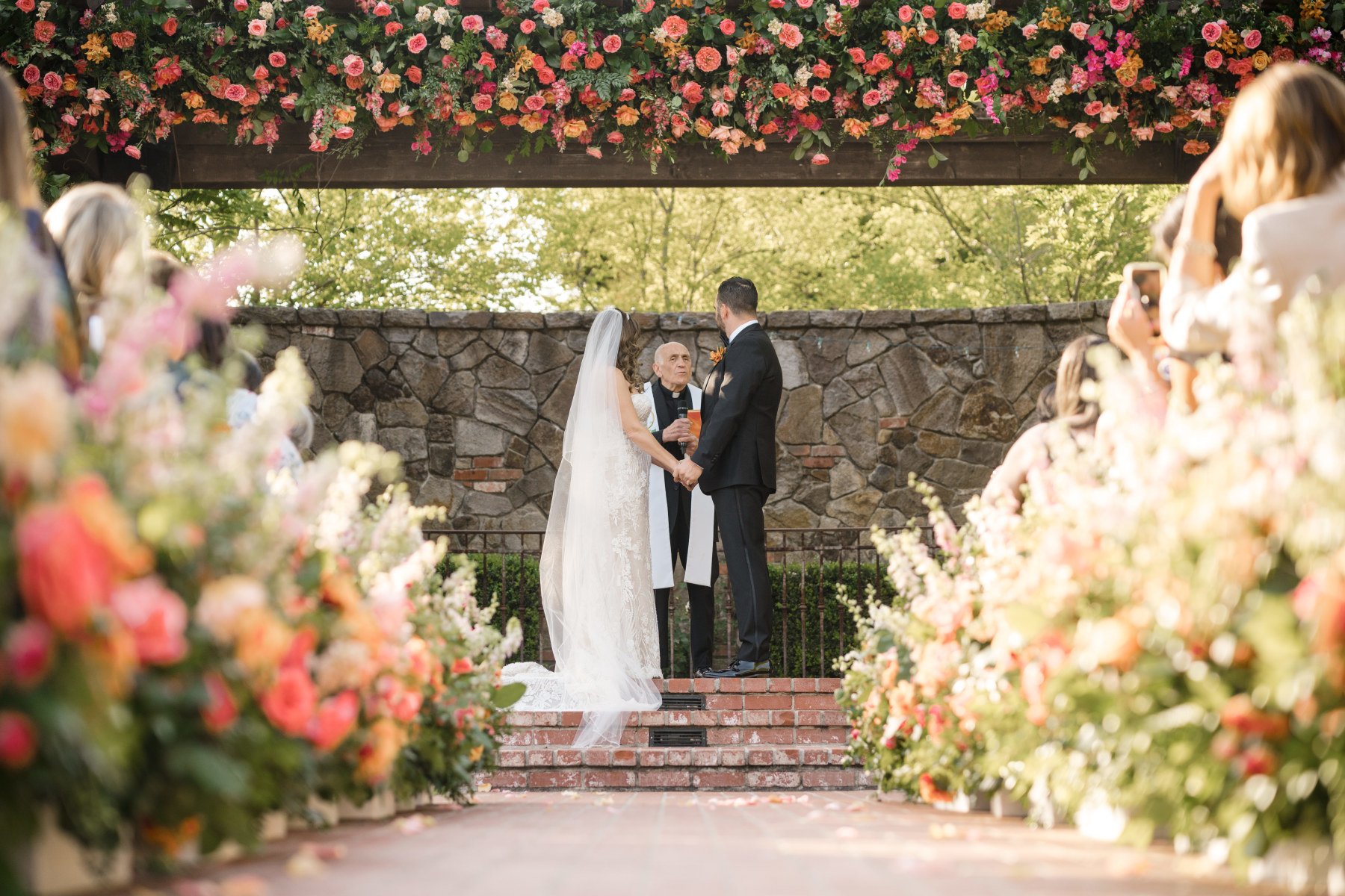 A couple getting married at the Yountville Wedding Venue are surrounded by an arch and a walkway  covered with roses and blooms.  Wedding planned by Blissful Events. Photo by Lily Rose. 