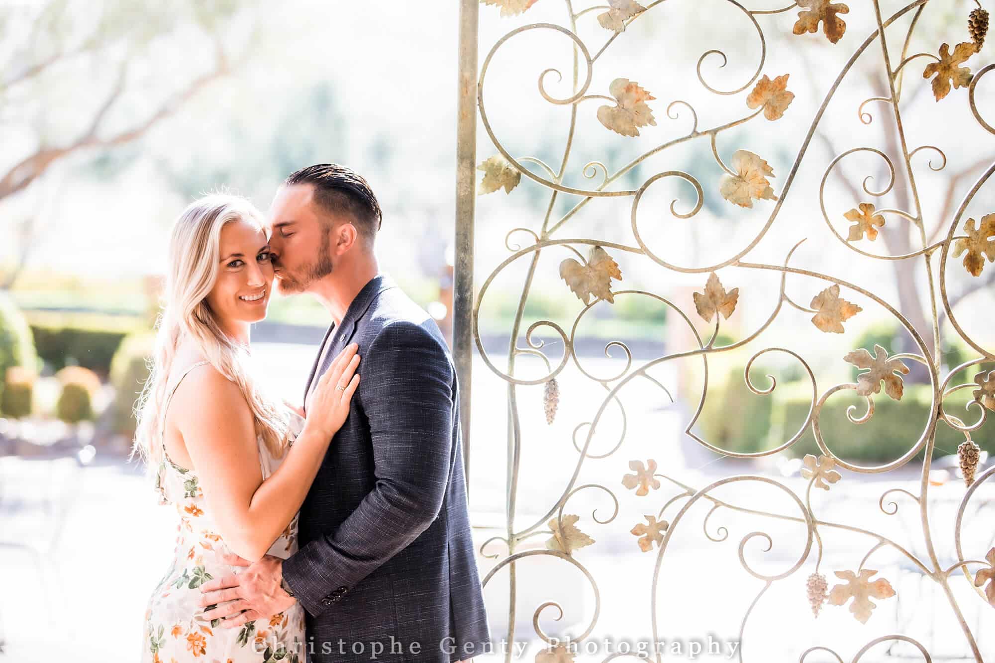 A newly engaged couple smile and kiss in front of an iron door shaped into vines at Viansa Winery a wonderful proposal location in wine country. Planned by Blissful Events. Photo by Christophe Gentry Photography.