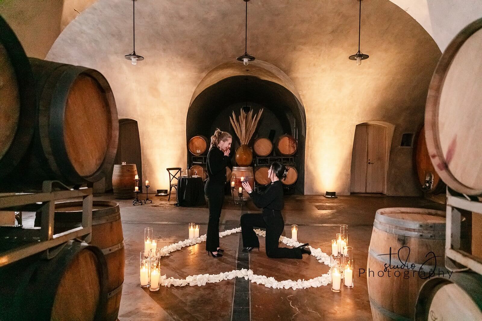 A woman proposes to her love in the Barrel Room of Viansa Winery surrounded by candles and white rose petals. Event planned by Blissful Events. Photo by Shiela Studio J Photography.