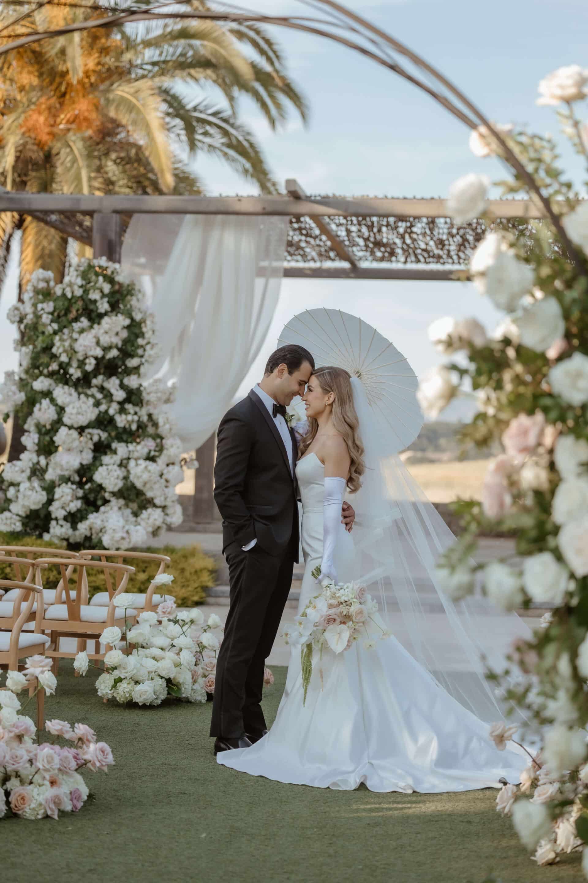 Bella Vita Floral Productions create breath-taking wedding flowers arrangements. A bride and groom stand in an archway of white flowers with bouquets lining the isle and outdoor stage. Photo by Andrea Lowry Photography. Wedding by Blissful Events.