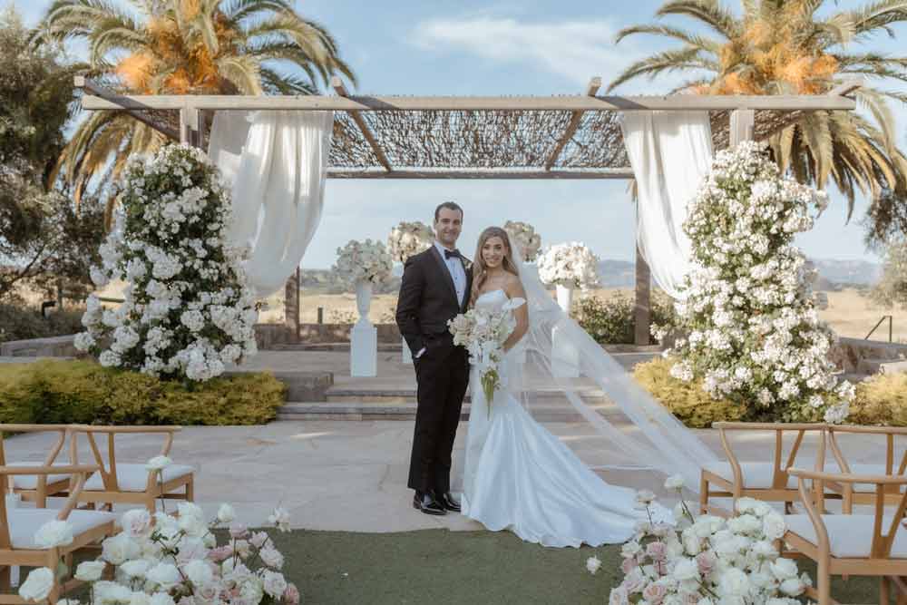Bella Vita Floral Productions create breath-taking wedding flowers arrangements. A bride and groom stand in an archway of white flowers with bouquets lining the isle and outdoor stage. Photo by Andrea Lowry Photography. Wedding by Blissful Events.