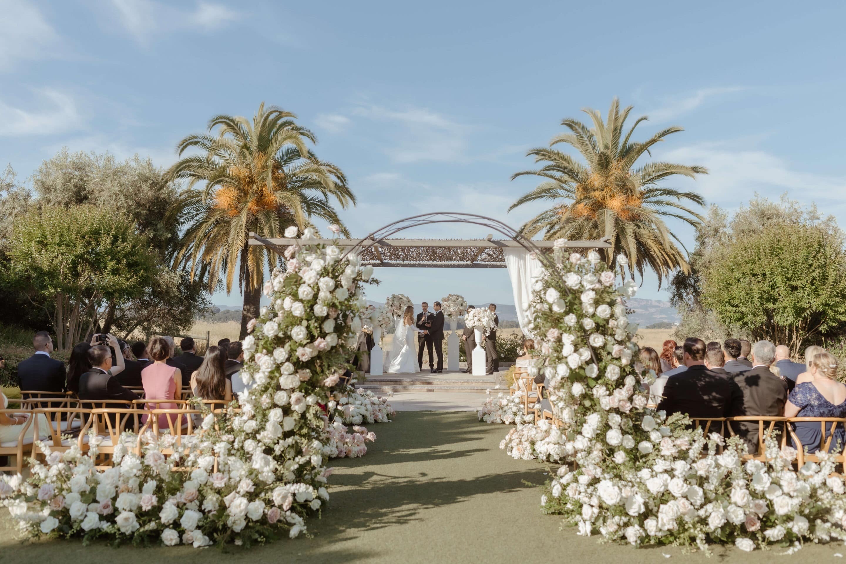 Bella Vita Floral Productions created the gorgeous floral arrangements surrounding the wedding ceremony. A bride and groom stand in an archway of white flowers with bouquets lining the isle and outdoor stage. Photo by Andrea Lowry Photography. Wedding by Blissful Events.